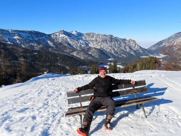 Mann sitzt schräg auf einer Bank im Schnee in der Wintersonne, im Hintergrund winterliche Berge 