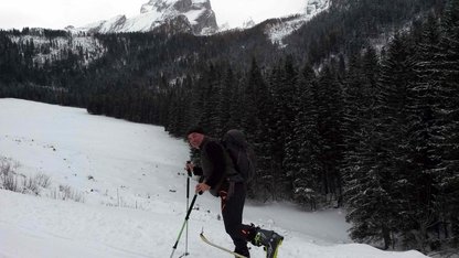 Lustiger Skibergsteiger auf einer winterlichen Waldlichtung, dahinter schroffe Berggipfel