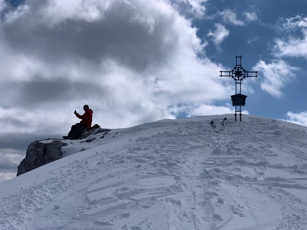Silhouette eines Skibergsteigers mit Daumen nach oben bei der Gipfelrast mit eisernen Gipfelkreuz 