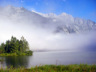 Bewaldete Halbinsel an Bergsee, dahinter Nebelschwaden unter Felsbergen 