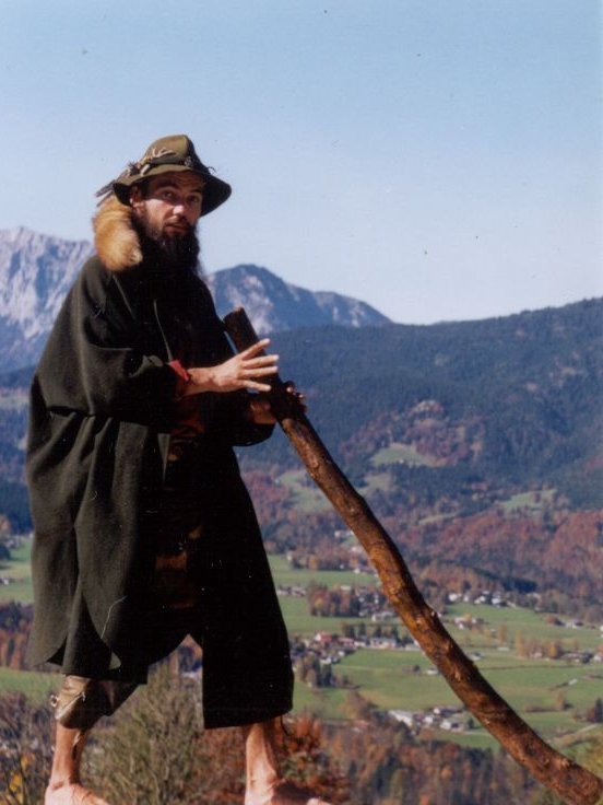 Mann mit langen Bart, grünen Hut mit Fuchsschwanz und langen grünen Mantel und Didgeridoo